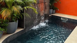 Ein Schwimmbecken mit Wasserfall an einer Wand, umgeben von Kübelpflanzen und einer orangefarbenen Wand.