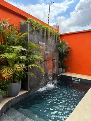 Una piscina con acqua che scende lungo una parete, circondata da piante in vaso e un muro arancione.