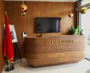A reception desk with a television above it, wooden paneling on the walls, and a red flag to the left. - HOTEL BLUE PEARL  (Dakhla)