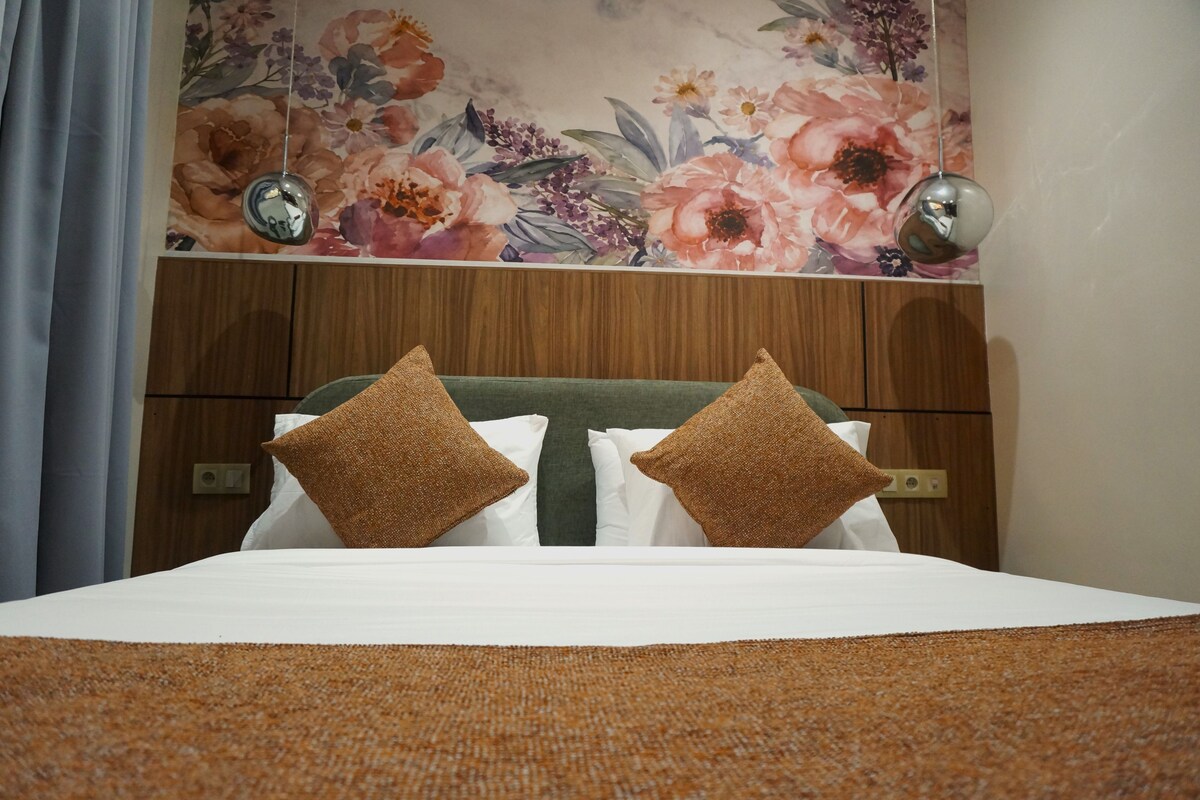 Ein Hotelzimmer mit einem Bett, zwei Kissen, einem hölzernen Kopfteil, einer Wanddekoration mit floralem Muster und einem Spiegel.