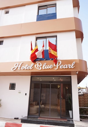 Entrada de un hotel con un cartel que dice 'Hotel Blue Pearl' y dos banderas sobre la entrada.