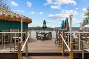 Een houten terras met buitenmeubilair, een rieten gebouw en uitzicht op een meer.