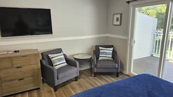 A room with a bed, two armchairs, a small round table, a TV mounted on the wall, and a sliding glass door leading to an outdoor area.