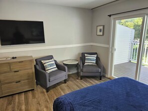 A room with a bed, two armchairs, a small round table, a TV mounted on the wall, and a sliding glass door leading to an outdoor area.