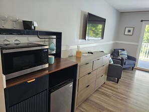 A modern kitchen with a microwave, refrigerator, and wooden countertop. There is a television mounted on the wall and a seating area with a sofa and armchairs.