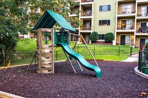 A playground with a slide and climbing structure, surrounded by a fence and greenery, with apartment buildings in the background. - Pigeon Forge | Romantic Mountain Retreat | Trails + Attractions (Pigeon Forge)