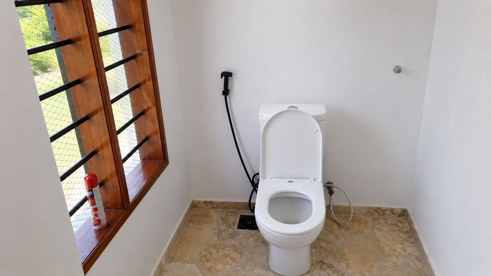 A small bathroom with a white toilet, a window with a wooden frame, and a tiled floor.