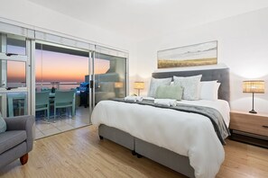 A modern hotel room with a large bed, a desk with a chair, a sofa, and a view of the sunset. - Sea Spray A109 (Cape Town)
