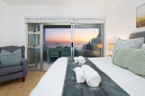 A hotel room with a bed, a sofa, a dining area with a table and chairs, and a view of the sea at sunset. - Sea Spray A109 (Cape Town)