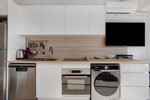A modern kitchen with white cabinetry, a built-in dishwasher, a stove, and a wall-mounted television. - Apartment 'Studio Plage et Village' with Sea View, Shared Pool and Air Conditioning (Le Barcarès)