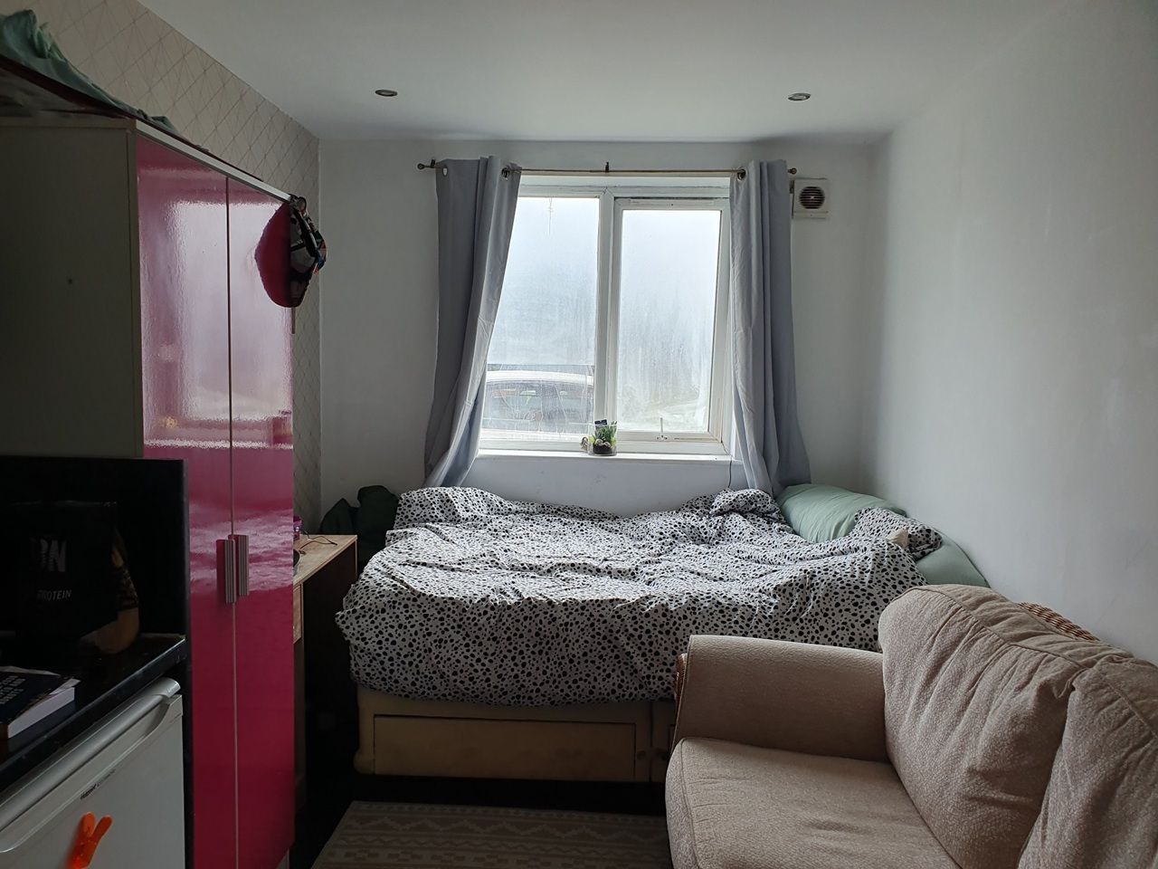 A small, compact room with a bed, a sofa, a kitchenette, and a window with curtains.