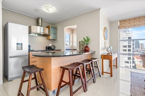 Eine moderne Küche mit einer zentralen Kochinsel, Barhocker, einem Edelstahlkühlschrank und Blick auf die Stadt durch große Fenster.