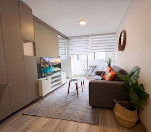 A modern living room with a flat-screen TV, a grey sofa, a glass coffee table, and a large plant. - Vista Bulnes Santiago by Domus Travels (Santiago)