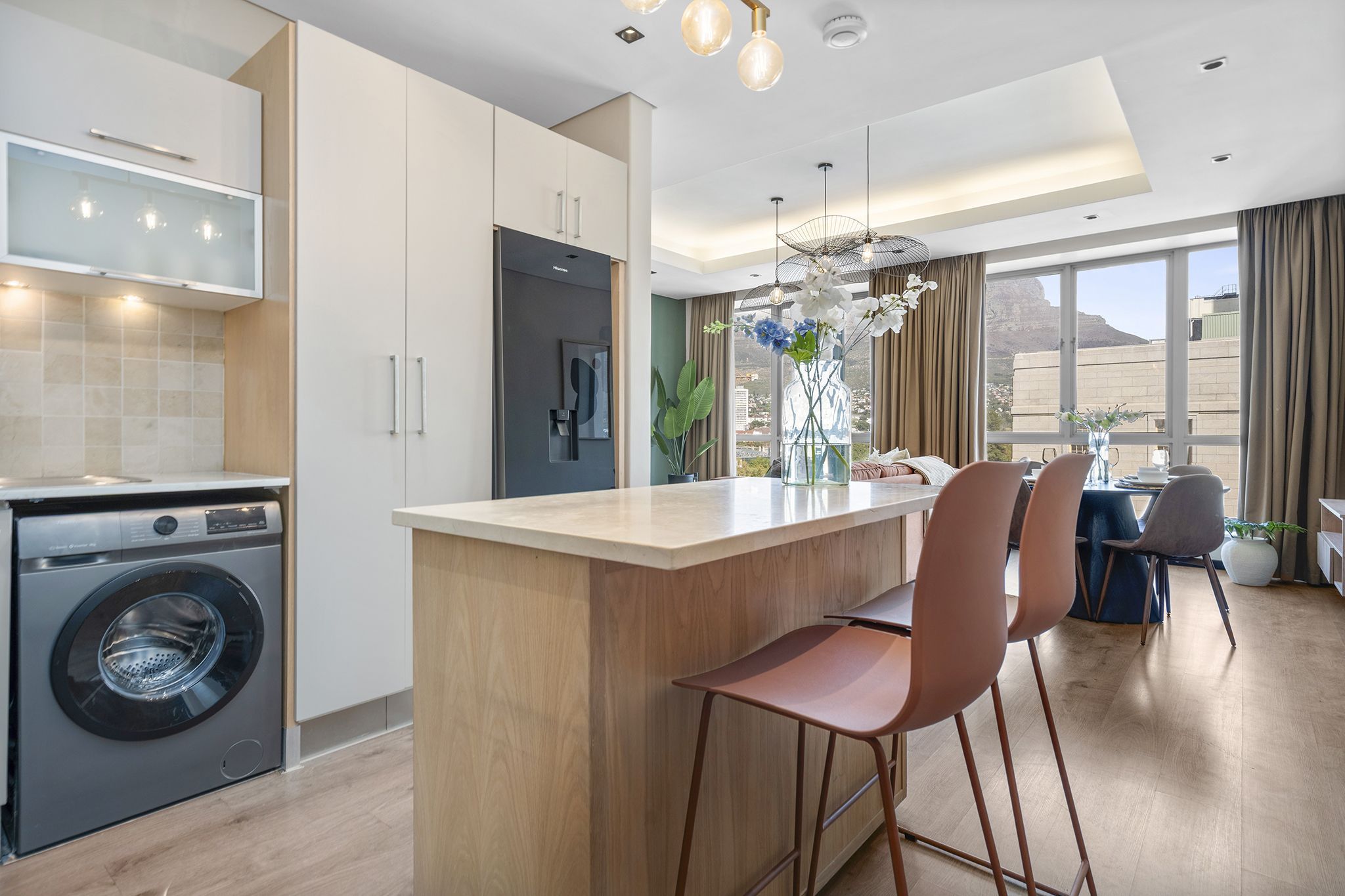 A modern kitchen with a built-in washer, a central island with bar stools, and a dining area with a view.