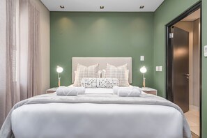 Ein ordentlich bezogenes Bett mit weißen Bettwäsche und Kissen, zwei Nachttischlampen und eine grüne Wand mit einem strukturierten Kunstwerk darüber.