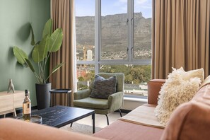 Sala de estar con vistas a un paisaje urbano y montañas, un sillón verde, una mesita y una planta en maceta.