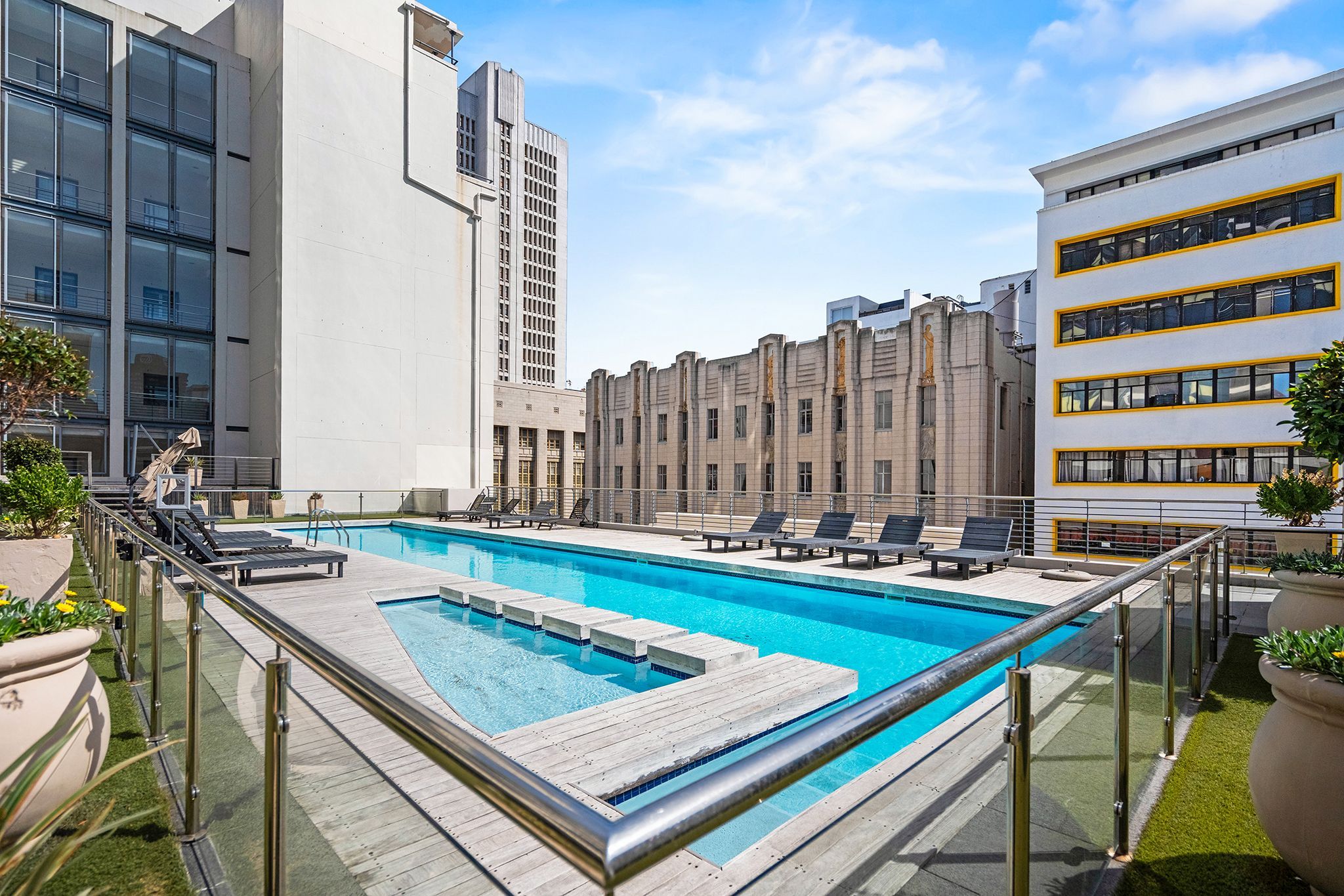 A rooftop pool with lounge chairs and a glass railing, surrounded by modern buildings.