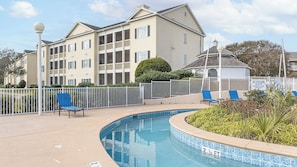 A swimming pool with a tiled edge, a white fence, and a building with balconies in the background. - Myrtle Beach | Poolside Leisure Break | Lazy River + Pool | 2BR (Myrtle Beach)