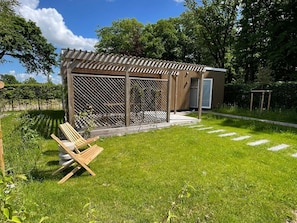 Eine kleine Holzkonstruktion mit überdachter Terrasse, einem umzäunten Garten und einer Bank.