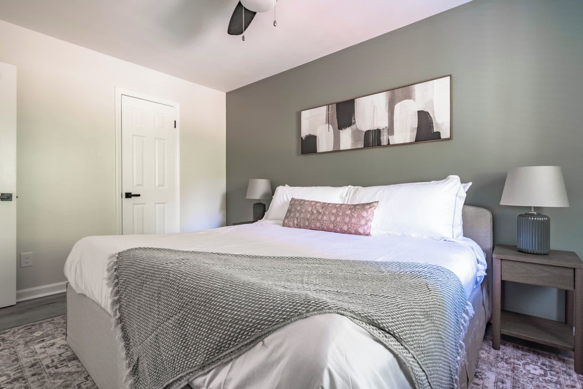 A bedroom with a bed, two bedside lamps, a door, and a wall-mounted abstract painting.