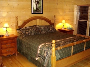 Ein hölzernes Bett mit Kopfteil, zwei Nachttische mit Lampen, ein gerahmtes Bild an der Wand und ein Fenster mit Jalousien.