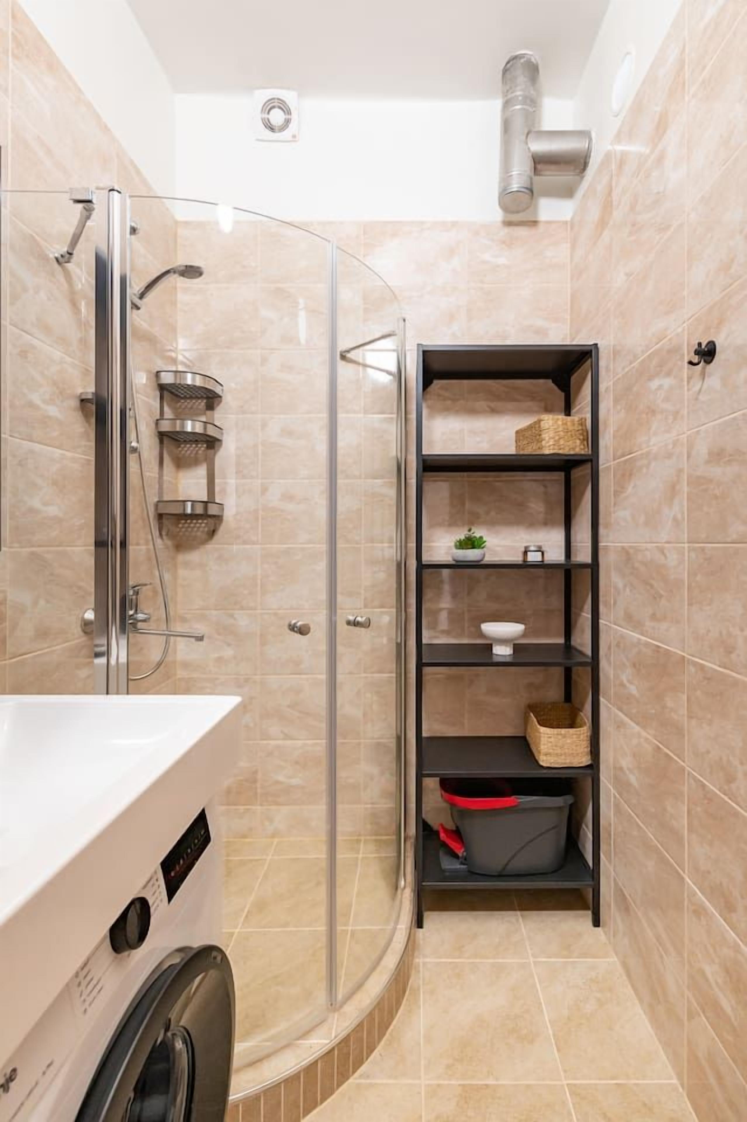 Un baño pequeño con ducha de cristal, lavadora integrada y estante con cestas.