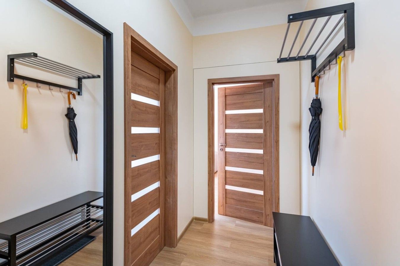 Un corridoio con porte in legno, un portariviste metallico con ombrelli e una panca nera.