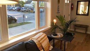 Ein Zimmer mit einer Couch, einem Tisch mit Lampe, einer Topfpflanze und einem Fenster mit Blick auf geparkte Autos und ein Gebäude.