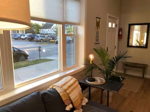 Una habitación con sofá, una mesa con lámpara, planta en maceta y ventana con vistas a coches aparcados y un edificio.
