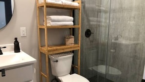 Ein Badezimmer mit Toilette, einem hölzernen Ablagebrett mit Handtüchern, einer Duschkabine aus Glas und einem weißen Waschbecken.
