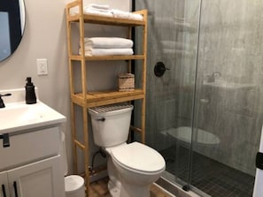 Un baño con inodoro, estante de madera con toallas, cabina de ducha de vidrio y lavamanos blanco.