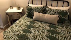 Ein Bett mit gemusterten Betttextilien, ein Nachttisch mit Lampe und ein an der Wand hängendes Bild.