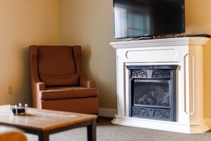 A living room with a fireplace, a television mounted above it, and a patterned armchair. - Lincoln NH | Mountain Escape | Pool, Hiking & Scenic Views | 2BR Suite (Lincoln)