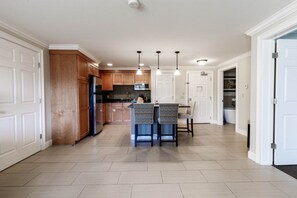A modern kitchen with wooden cabinets, a central island, pendant lighting, and a view into the dining area. - Lincoln NH | Mountain Escape | Pool, Hiking & Scenic Views | 2BR Suite (Lincoln)