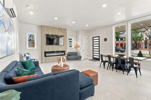 A modern living room with a sofa, a fireplace, a dining area with chairs, and a large window offering a view of the street. - Modern 3BR Home Near Midtown & Atlantic Station (Atlanta)