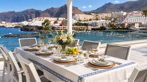 An outdoor dining table set with plates, glasses, and a fruit bowl, overlooking a coastal town and mountains. - Luxury Villa with a Heated Pool and Panoramic Ocean Views (Santiago del Teide)