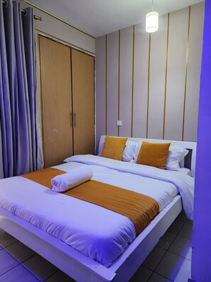 A bed with white and orange bedding, a wooden wardrobe, and a hanging light fixture. - 
Cozy Modern Studio on Ngong Road (Nairobi)