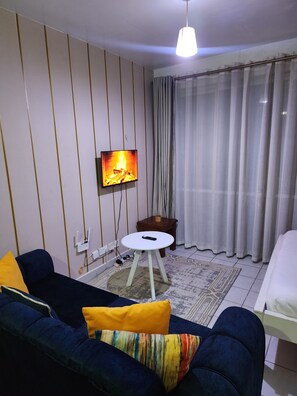 Quarto de hotel com sofá, mesa redonda pequena, cama, televisão fixada na parede e janela com cortinas.