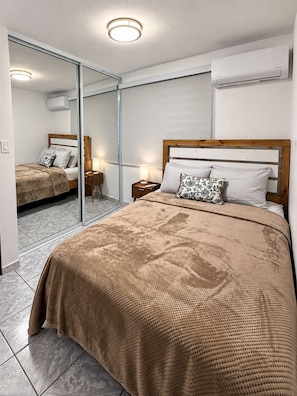 Hotellihuone, jossa on suuri sänky, yöpöytä lampun kanssa, peili ja ilmastointilaite seinässä.