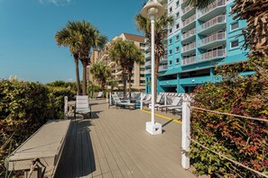 A wooden deck with lounge chairs, a palm tree, and a building with balconies. - North Myrtle Beach | Beach Escape | Pool & Ocean Access | 1BR Villa (North Myrtle Beach)