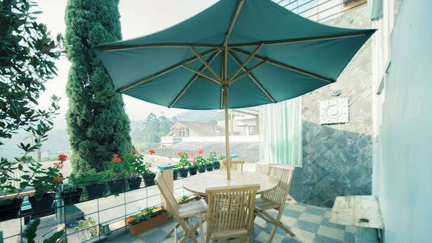 Een terras met een tafel en stoelen, een grote parasol, bloempotten en een uitzicht op een gebouw in de verte.
