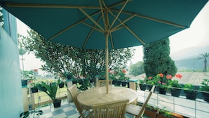 Een ronde houten tafel met vier stoelen en een grote parasol, omringd door potplanten en bloemen op een balkon met een geruite vloer.