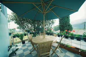 A round wooden table with four chairs and a large umbrella, surrounded by potted plants and flowers on a balcony with a checkered floor. - Rehat at Villa Cemara Kembar (Kabupaten Bandung)