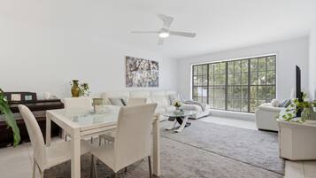 Uma sala de estar moderna com uma mesa de jantar de vidro, sofás brancos, uma janela grande com persianas e um ventilador de teto.