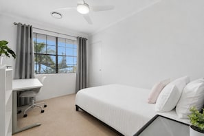 Quarto de hotel com cama, escrivaninha, cadeira, ventilador de teto e janela com cortinas.
