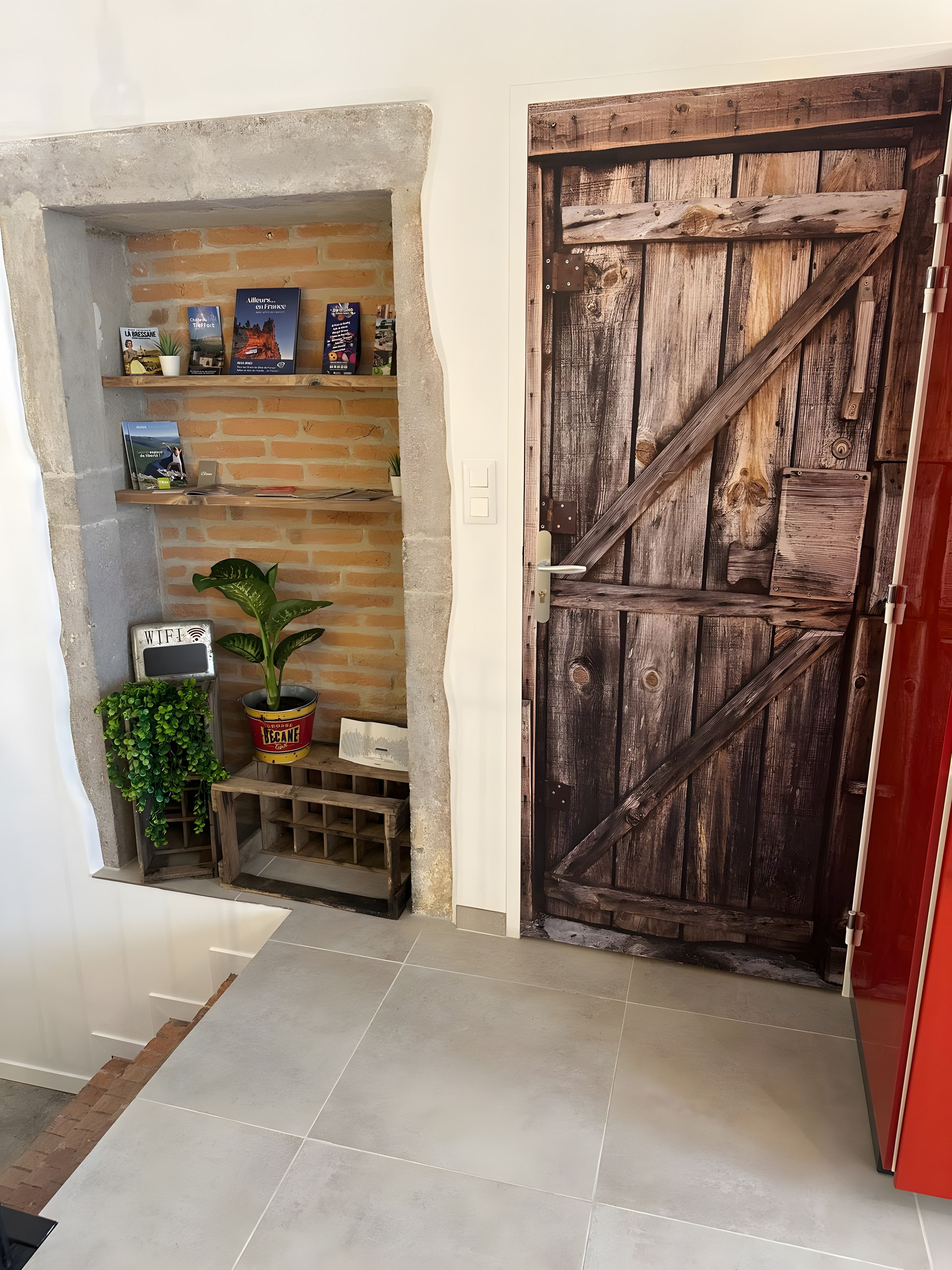 Un intérieur rustique avec une porte en bois, un mur en briques et une étagère où sont posés des livres et un pot de fleurs.