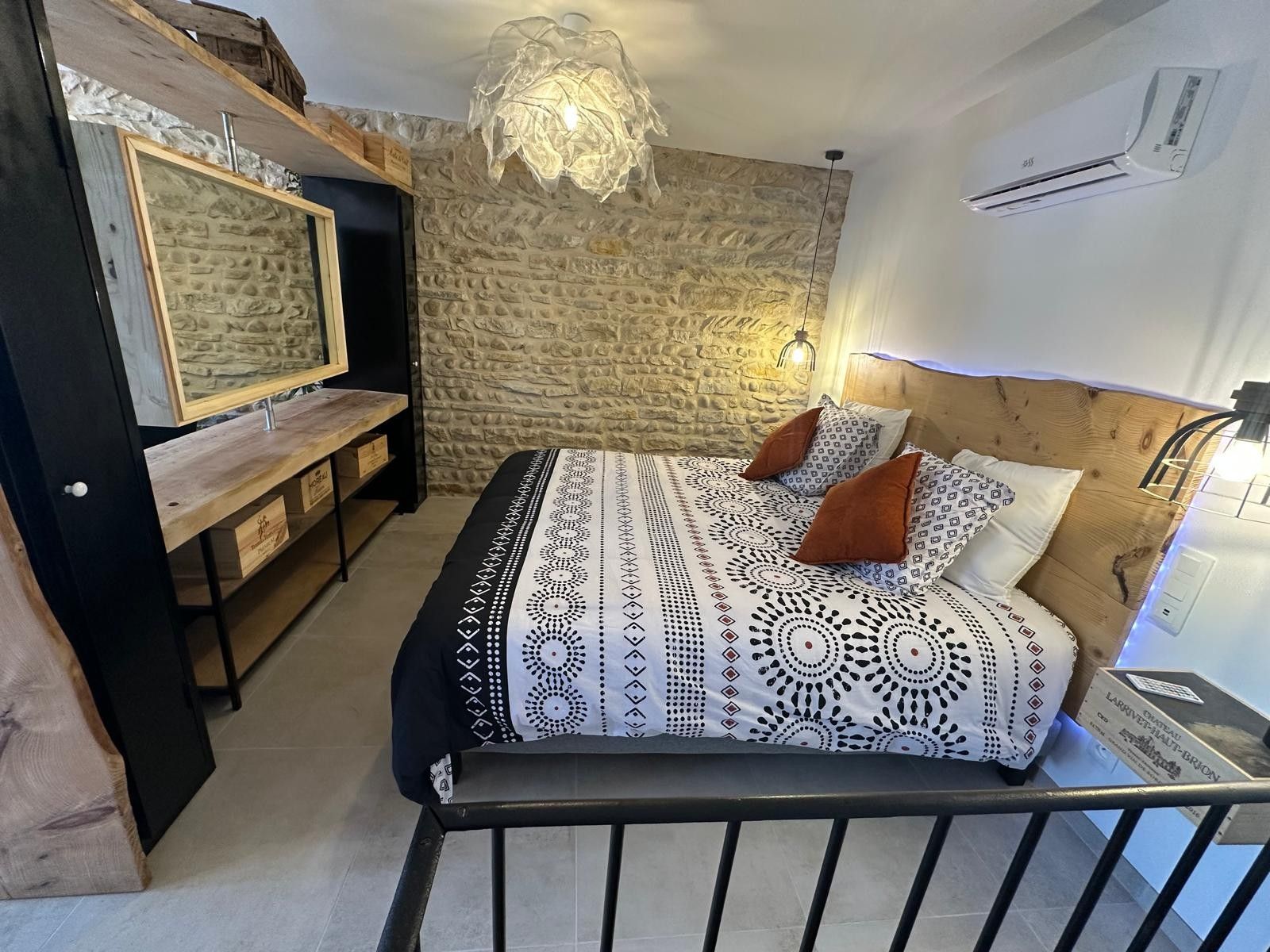 Une chambre avec un mur en pierre, un lit avec une literie à motifs, une tête de lit en bois, un téléviseur fixé au mur et un luminaire au plafond.