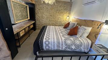 Une chambre avec un mur en pierre, un lit avec une literie à motifs, une tête de lit en bois, un téléviseur fixé au mur et un luminaire au plafond.