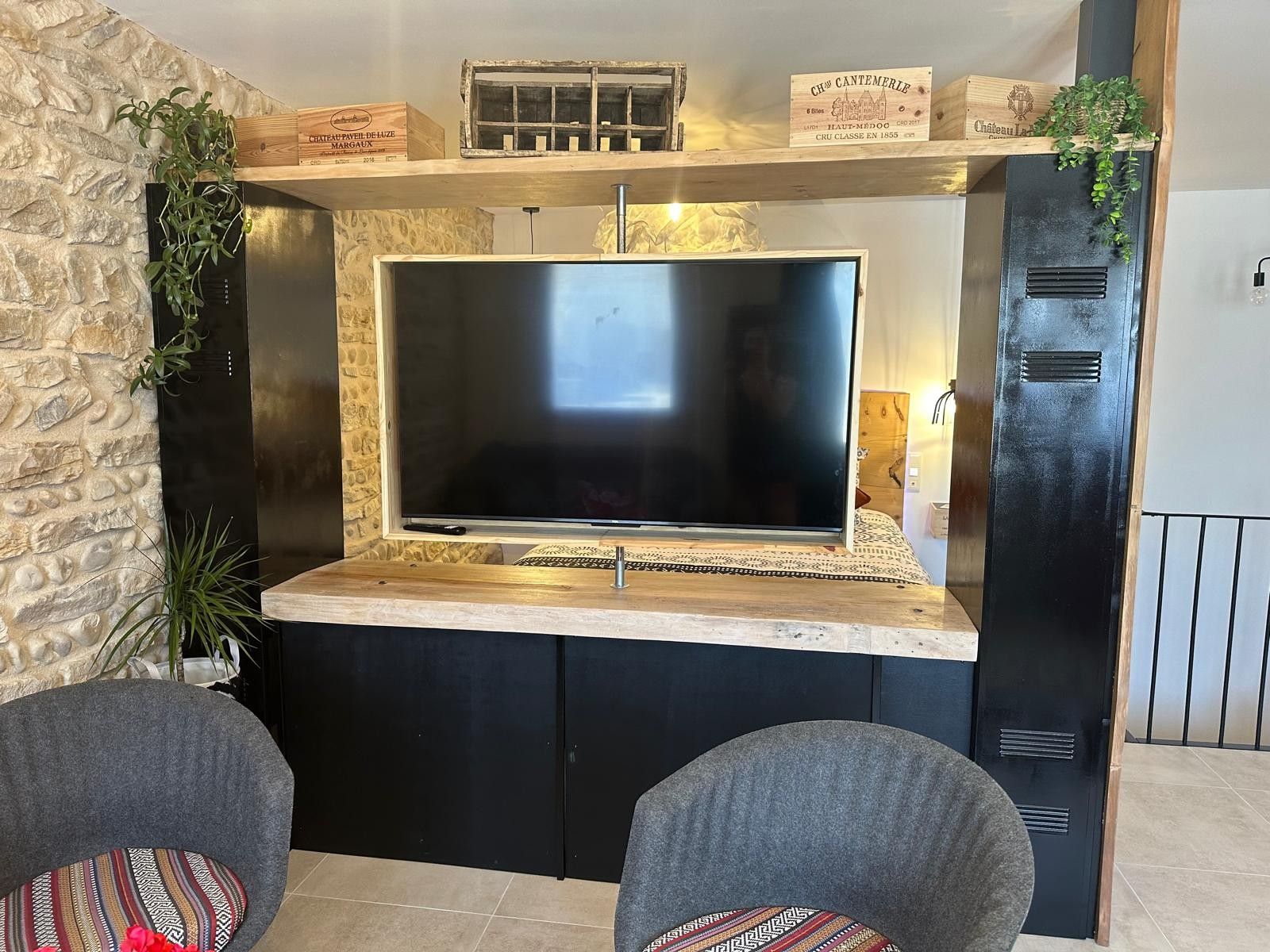 A modern living area with a large flat-screen TV mounted on a stone wall, a wooden shelf with decorative items, and a black cabinet with ventilation grilles.