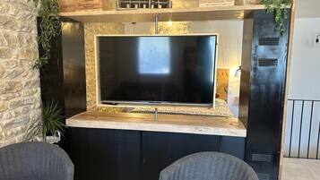 A modern living area with a large flat-screen TV mounted on a stone wall, a wooden shelf with decorative items, and a black cabinet with ventilation grilles.